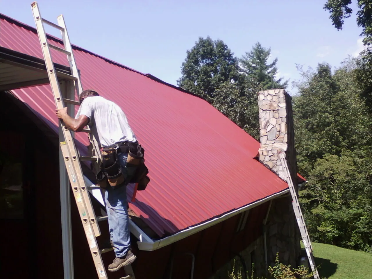 Roofer on a ladder beside a red metal roof and stone chimney — Soffit & Fascia Repair by Roofing Brothers in Stockton